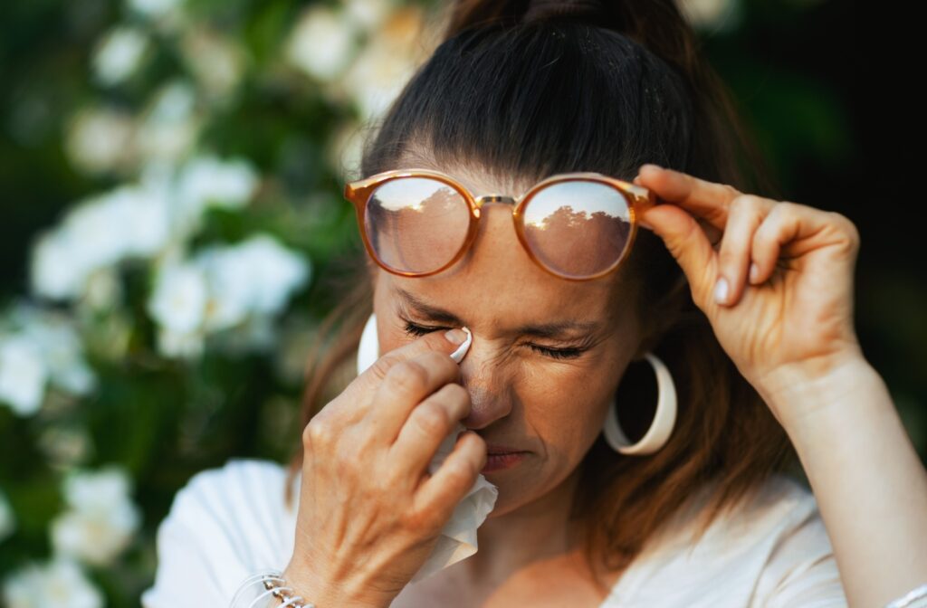 Someone with eye allergies lifts their glasses and rubs their eye with a tissue.