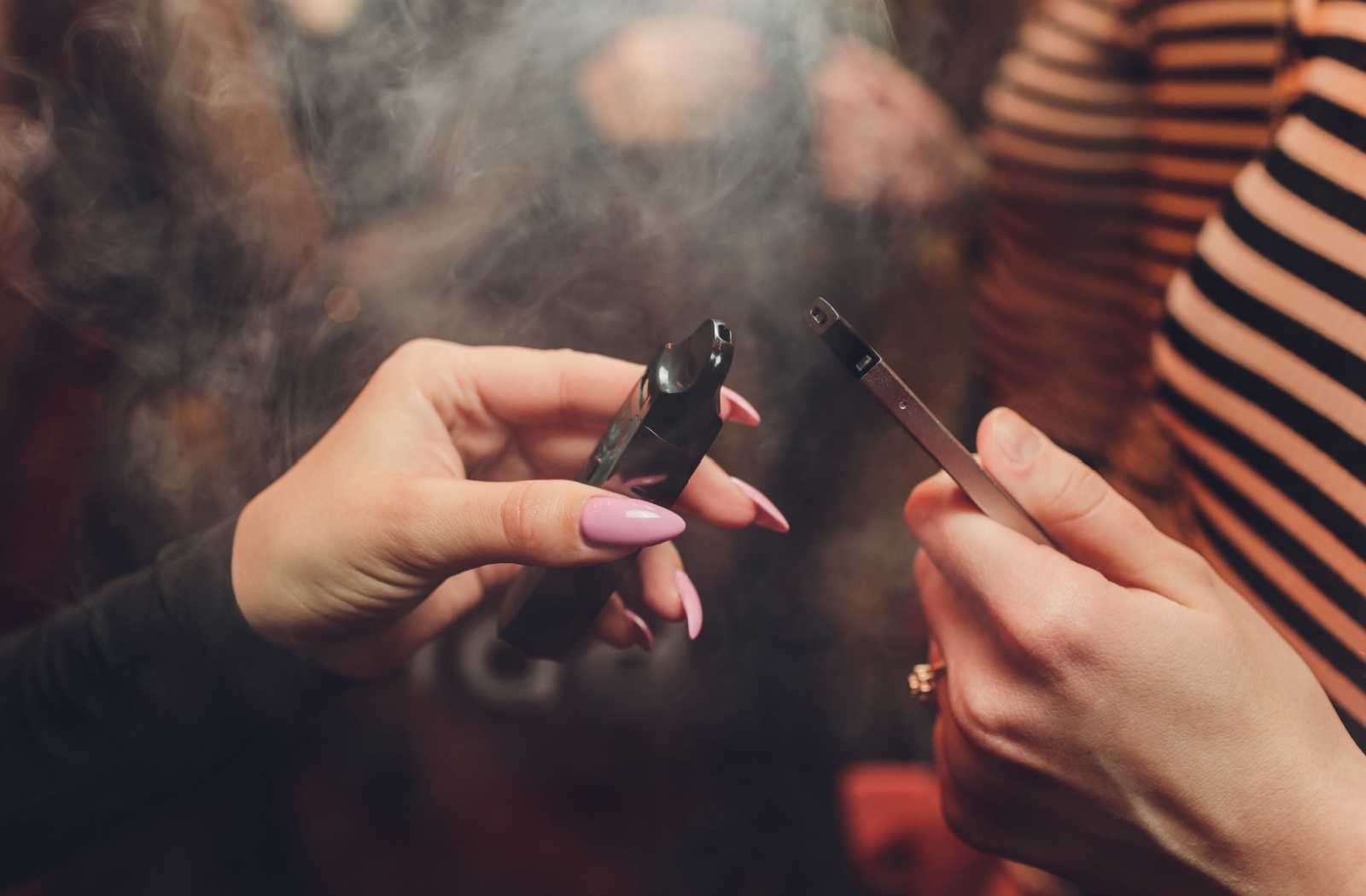 Two hands holding two vape pens with smoke surrounding.